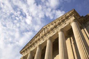 A low-angle view of a neoclassical government building with tall, ornate columns and a detailed pediment under a partly cloudy sky. The structure is bathed in warm sunlight.
