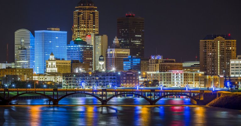 Nighttime view of a city skyline with modern high-rise buildings and historic architecture, showcasing one of the best states. The buildings are illuminated, reflecting on the calm river below. A bridge with arched supports spans the river, also lit with colorful lights.
