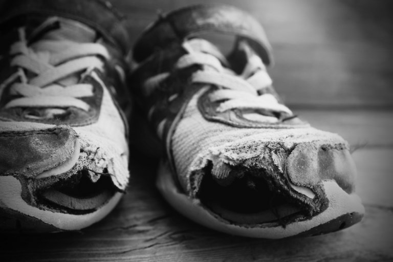 A grayscale image of a pair of worn-out sneakers with frayed edges and holes in the front. The shoes are placed side by side on a wooden surface, capturing the texture and extensive wear, reminiscent of countless hours spent in various work programs.