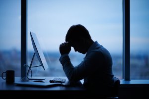 Person sitting at a desk in a dimly lit room, with head resting on hand, clearly showing signs of stress. A desktop computer, keyboard, and coffee mug are on the desk. The large window in the background reveals a blurred cityscape at dusk.