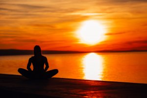 A person sits cross-legged facing a calm body of water, silhouetted against a vibrant sunset. The sky is ablaze with shades of orange, red, and yellow as the sun descends towards the horizon, casting reflections on the water's surface—a perfect escape from stress.