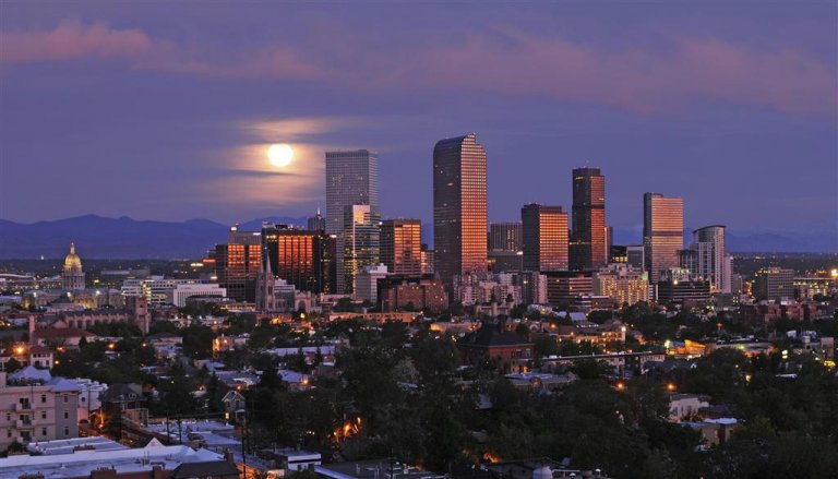 A view of Denver's skyline at dusk, with the full moon rising behind the buildings. The skyscrapers are partially illuminated by the fading sunlight, casting a soft glow over Mile-High City. The distant mountains and a mix of trees and buildings are visible in the foreground.