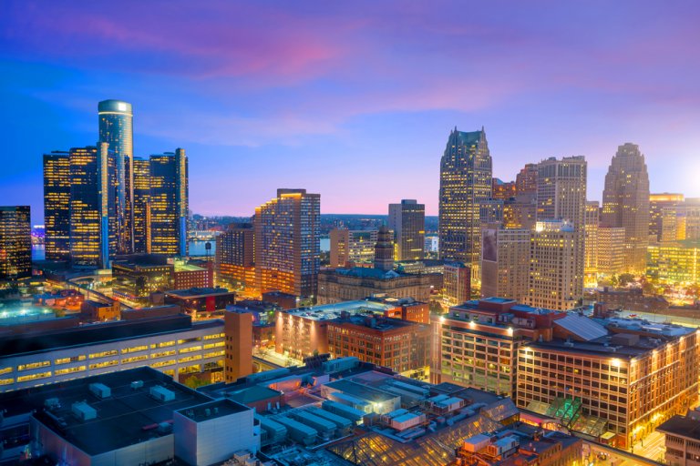 A vibrant cityscape of downtown Detroit at dusk, showcasing a mix of illuminated skyscrapers and buildings against a stunning twilight sky. The iconic Renaissance Center is prominently featured among the skyline, surrounded by a blend of modern and historic architecture.