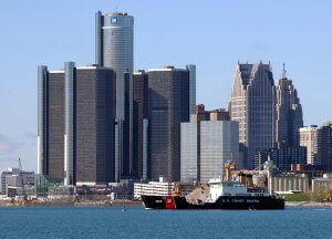 A U.S. Coast Guard ship sails on the river with the Detroit skyline in the background. Dominating the skyline is the Renaissance Center, a cluster of modern skyscrapers. Classic older buildings are also visible, contrasting with the modern architecture.