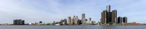 Panoramic view of a city skyline across a body of water. Tall skyscrapers dominate the horizon with a mix of modern and older buildings. The sky is mostly clear with a few clouds. The water in the foreground appears calm, reflecting part of the skyline.