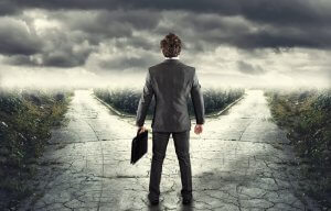 A man in a suit stands at a fork in the road, holding a briefcase. The sky is overcast with dark, menacing clouds. The road ahead splits into two paths, symbolizing his career choices as they disappear into the horizon amidst a vast, desolate landscape.