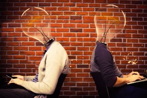 Two individuals sit back-to-back in front of laptops, each with a light bulb instead of a head. The filament of each bulb is connected by a wire, symbolizing the shared thinking essential for career growth. They are positioned against a brick wall background.
