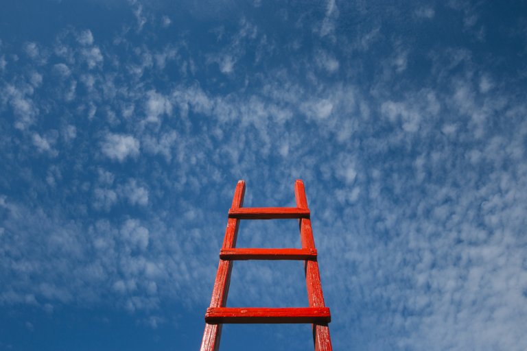 A bright red ladder extends upward against a backdrop of a vibrant blue sky dotted with scattered white clouds. The ladder appears to be reaching towards the sky, conveying a sense of ambition, much like the pursuit of new jobs.