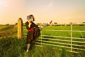 A person in a black dress and striped leggings performs a high kick while holding a book in front of their face. They are standing on a grassy field next to a metal gate and a wooden post, with the sun shining brightly in the background, blending athleticism with literary culture.