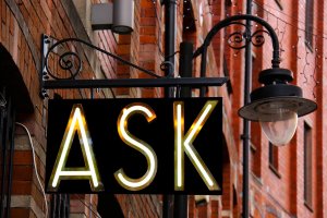 A black rectangular sign with the word "ASK" in yellow neon letters hangs from an ornate metal bracket. The sign, symbolizing leadership, is attached to a red brick building adorned with string lights along the overhang and an old-fashioned street lamp nearby.