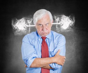 An older man with white hair and a serious expression stands with his arms crossed, exuding leadership. He is wearing a light blue dress shirt and a red tie. A steam-like effect appears to come out of his ears against a dark, textured background.