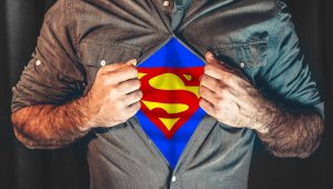 A person is pulling open their shirt to reveal a blue undershirt with a large yellow and red Superman logo underneath, symbolizing leadership. The person’s head is not visible in the image, and the background is dark.