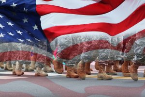 The image shows soldiers in camouflage uniforms and brown boots marching in unison with an American flag overlaying the scene. The soldiers' legs are visible, and the prominent stars and stripes of the flag add a patriotic effect, symbolizing their dedication and sacrifice for the nation.