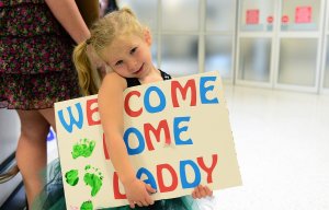 A young girl with blonde pigtails and a colorful dress holds a handmade sign that reads "Welcome Home Daddy" in blue and red letters. Green handprints are painted on the sign. She stands in an airport, smiling shyly with her head tilted, honoring her father's sacrifice.