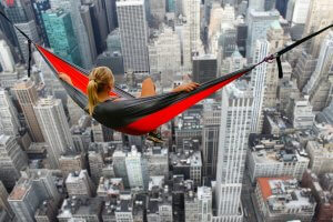 A woman is lounging in a red and black hammock suspended high above a cityscape filled with skyscrapers, giving a dramatic and breathtaking view of the buildings below. As she gazes towards the horizon, her serene demeanor seems to subtly promote her personal brand of adventurous elegance.