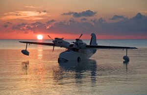 A seaplane floats on calm water during sunset. The sky, filled with orange and pink hues as the sun touches the horizon, creates a breathtaking backdrop for admiring seaplanes. Clouds partially cover the sky, reflecting the warm colors of the setting sun.