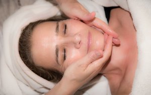 Close-up of a person with closed eyes receiving a facial massage. Their head is wrapped in a white towel, and the hands of the masseuse are gently massaging their cheeks and jaw. The professional image captures the relaxed and content expression perfectly.