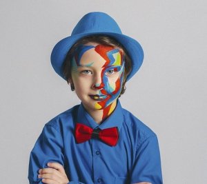 A child wearing a blue shirt and a blue hat, with a red bowtie, poses for a professional image. The child has vibrant, multicolored face paint creating an abstract design covering one side of their face. The background is plain and light-colored.