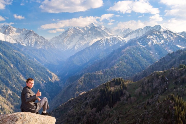 A person wearing a gray jacket sits on a large rock overlooking a vast mountain range with snow-capped peaks and lush forests under a partly cloudy sky, giving a thumbs-up gesture, as if celebrating the natural succession of seasons.