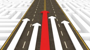 An illustration of a road with five arrows pointing forward. The middle arrow is red and larger than the four white arrows flanking it, which are all of equal size, symbolizing succession. The background contains a maze pattern, representing direction and progress.