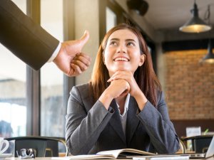 A woman in a business suit is sitting at a table, smiling and looking up, with her hands resting on an open book. A person standing next to her is giving her a thumbs up, silently conveying their thanks. The background shows a modern office space with large windows and brick walls.