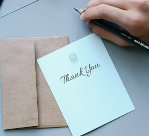 Close-up of a person holding a pencil and writing on a light blue card with "Thank You" written in elegant cursive. Underneath the card is a brown envelope, and a second envelope is visible in the background on the table, ready to be sent to those being thanked.