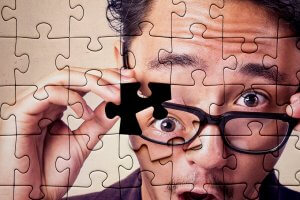 A close-up of a person wearing glasses, with the image designed to look like a jigsaw puzzle. They are holding one puzzle piece in front of their face, revealing part of their eye. The person's expression appears surprised or intrigued, evoking a sense of employee engagement and discovery.
