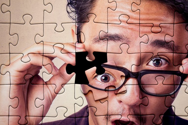 A close-up of a person wearing glasses, with the image designed to look like a jigsaw puzzle. They are holding one puzzle piece in front of their face, revealing part of their eye. The person's expression appears surprised or intrigued, evoking a sense of employee engagement and discovery.