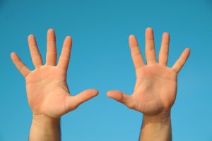 Two hands with open palms facing the camera are raised against a clear blue sky. The two hands appear to be from the same person, positioned symmetrically and fully extended.