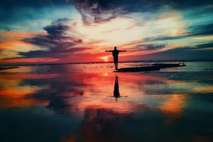Silhouette of a person standing with two hands outstretched on a beach during a vibrant sunset, reflected in the wet sand. The sky is painted with swirls of orange, pink, blue, and purple clouds, creating a serene and picturesque scene.