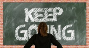 A person with medium-length brown hair, wearing a black jacket, stands facing a chalkboard with the words "KEEP GOING" written in large chalk letters, their two hands resting gently on the board. The chalkboard is framed by a brick wall.