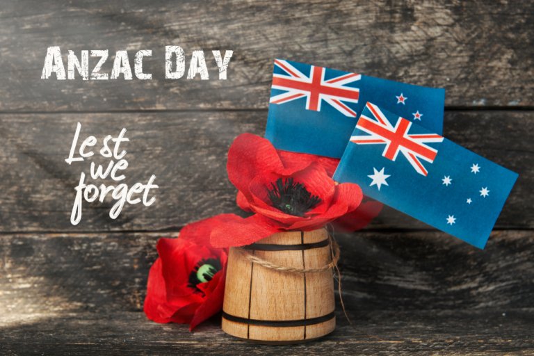 A rustic wooden background with the words "ANZAC DAY" and "Lest we forget" in white text, accompanied by two small Australian flags and red poppies in a wooden barrel, symbolizing remembrance and honor for fallen Anzac soldiers.