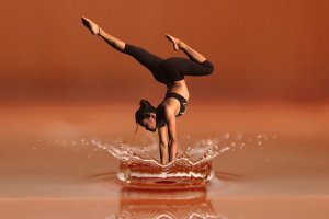 A woman in athletic wear performs a handstand with legs split, balancing on one hand. She appears to be frozen mid-air above a rippling splash of water that reflects her image. The background resembles the rich, warm hues often associated with ANZAC commemorations.