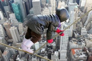 A child wearing a puffy coat, pink gloves, and boots is balancing on a tightrope high above an Anzac memorial in a city skyline filled with tall buildings. The child is crouching and holding onto the rope for support. The background shows a cityscape from an aerial perspective.