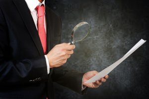 A person in a black suit and red tie uses a magnifying glass to examine a document, ensuring compliance. The background is dark and abstract, highlighting the focus on the detailed inspection of the paperwork.