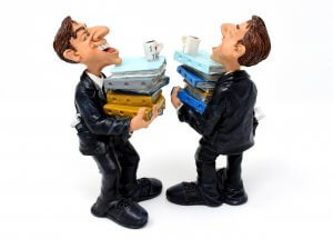 Two cartoonish figurines dressed as office workers in black suits and ties are standing face-to-face, each holding stacks of binders of various colors. Both have exaggerated happy expressions, and each stack has a coffee cup on top. Their jovial demeanor adds a fun touch to the world of compliance.