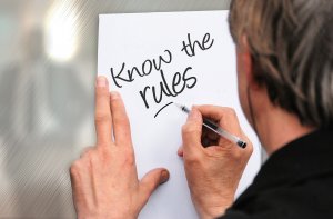 A person with gray hair is holding a white sheet of paper and writing "Know the rules" in black marker, emphasizing the importance of compliance. Their left hand steadies the paper while their right hand writes. The background is blurred and features a metallic texture.