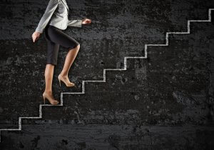 A person in business attire, wearing a gray blazer, black pants, and beige high heels, is climbing a set of white stairs against a dark, textured background. The image captures the lower half of their body, emphasizing their upward movement in what symbolizes career development.