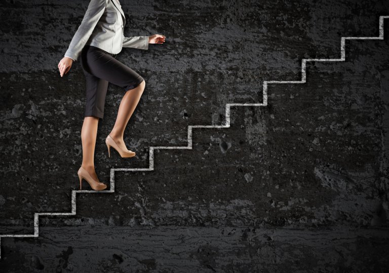 A person in business attire, wearing a gray blazer, black pants, and beige high heels, is climbing a set of white stairs against a dark, textured background. The image captures the lower half of their body, emphasizing their upward movement in what symbolizes career development.