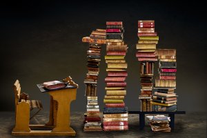 An old wooden desk and chair sit to the left of three tall, uneven stacks of worn, vintage books on career development. To the right, a small table supports additional books, with more scattered on the ground. The background is dark and minimalist, highlighting the cluttered book arrangements.