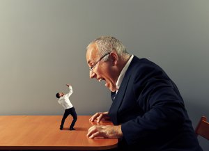 A composite image depicting a large man in a suit, appearing to shout criticism at a much smaller man who is standing on a table. The larger man is leaning forward with an angry expression, while the smaller man is leaning back in fear.