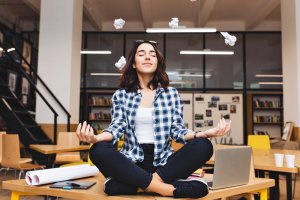 A woman in a plaid shirt sits cross-legged on a desk, meditating with eyes closed and hands in a zen position. Crumpled papers are suspended in mid-air around her like a chaotic template. There's a laptop, papers, and a pencil on the desk, with bookshelves in the background.