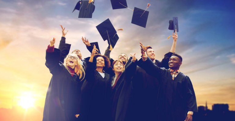 A group of graduates in black gowns and caps, soon-to-be employees, are joyfully tossing their caps into the air against a backdrop of a vivid sunset. They are smiling and looking upwards, celebrating their accomplishment.