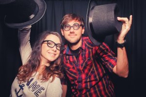 A smiling woman and man, both employees wearing glasses, hold up top hats in a playful manner. The woman has long brown hair and wears a gray shirt, while the man has short hair, a beard, and wears a red plaid shirt. Both are standing against a dark curtain backdrop.