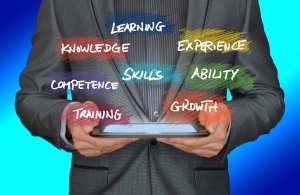 A person in a suit holding a tablet stands surrounded by colorful words: "LEARNING," "KNOWLEDGE," "EXPERIENCE," "SKILLS," "ABILITY," "COMPETENCE," "GROWTH," and "TRAINING." Each vibrant word represents key aspects of human resources, set against a blue background.