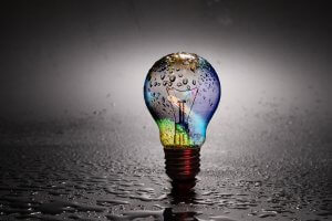 A light bulb with a colorful, iridescent surface is standing on a wet surface, covered in water droplets. The background is a gradient from dark to light gray, creating a reflective effect on the surface. Unlit, the bulb displays its intricate filament like an artifact of government engineering.