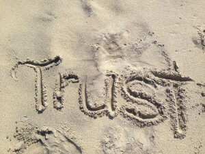 The word "Trust" is written in the sand with visible footprints around it, symbolizing loyalty and mutual respect. The sand is smooth and light-colored, with some shadows indicating slight unevenness in texture.