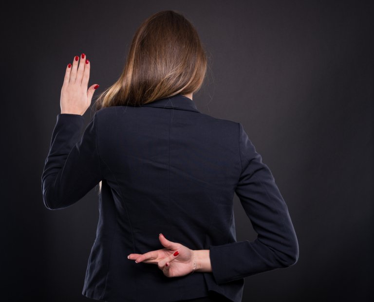 A woman with long brown hair, wearing a dark blazer, is shown from behind against a dark background. She has her right hand raised as if taking an oath of loyalty, while her left hand is behind her back with fingers crossed.