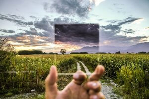A hand holds up a small tinted glass sheet against the background of an expansive field with a dirt path, lined with greenery and mountains in the distance under a dramatic sky with thick clouds and a sunset. The glass, loyal to its purpose, alters the colors of the scenery it frames.