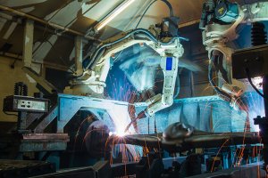 Two robotic arms are welding metal in an industrial setting, emitting bright blue light and sparks. The environment appears to be a factory or manufacturing plant with various machinery and equipment visible in the background, illustrating a crucial role in national economic development.